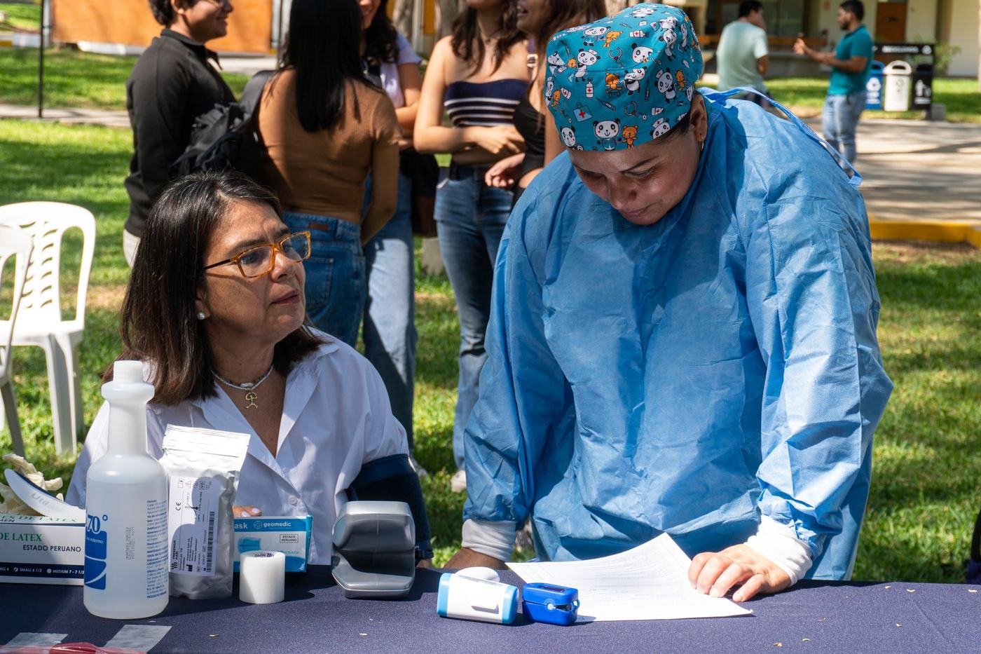 Una médica con cubrebocas y gorro escribe en una mesa de registro de una jornada de salud al aire libre; una mujer con lentes espera sentada al lado y otras personas hacen fila al fondo.