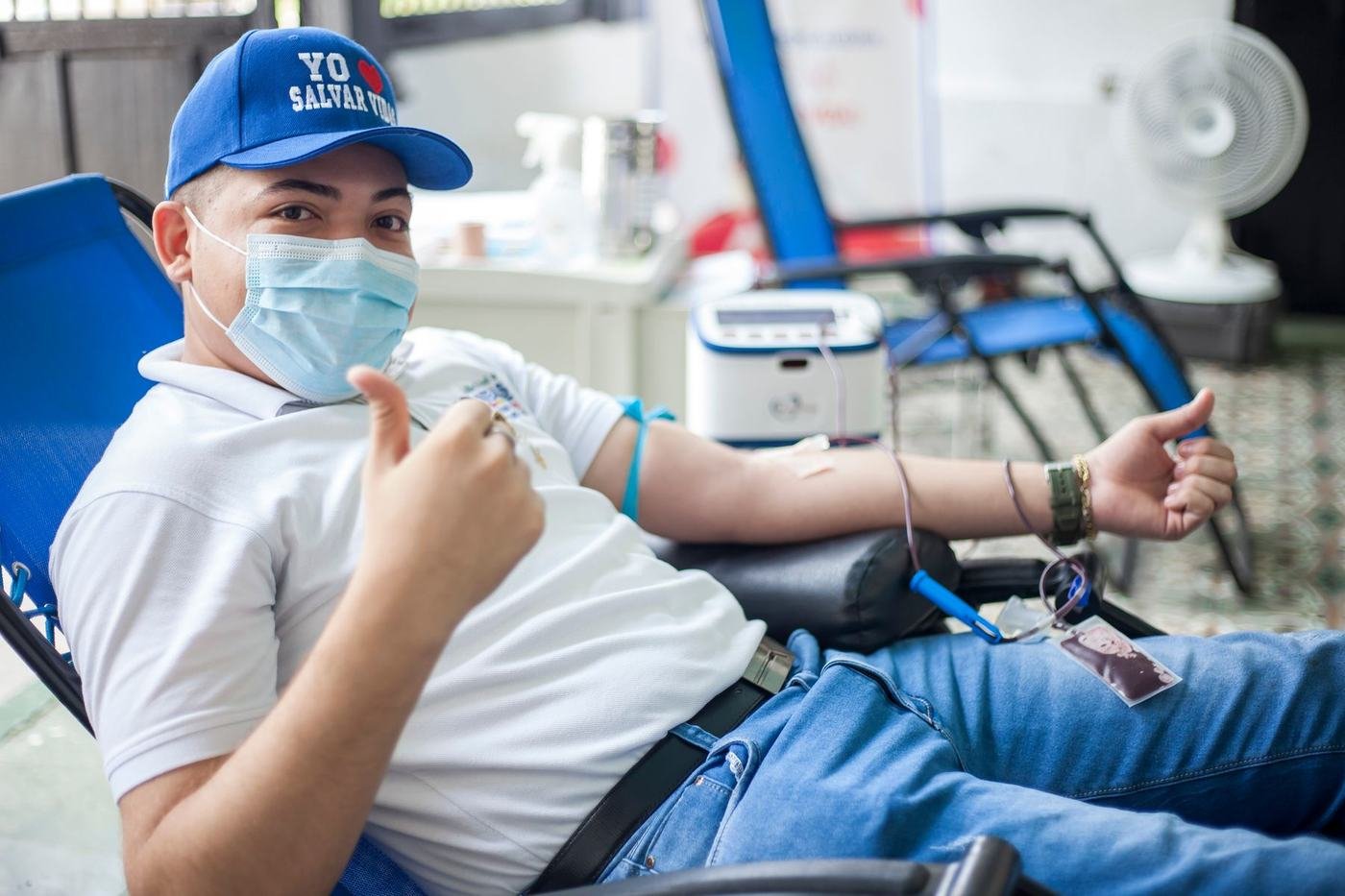 Un voluntario con gorra que dice Yo amo salvar vidas levanta el pulgar sonriendo durante una jornada de donación de sangre.